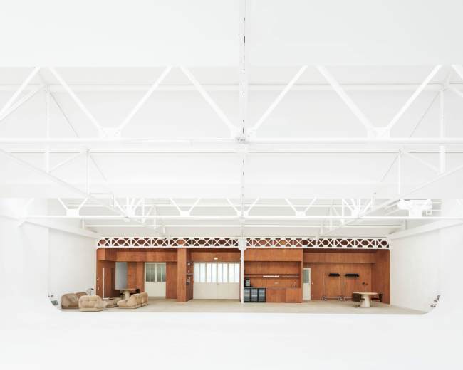 The image shows a spacious, minimalist interior featuring wooden cabinetry, a couple of beige sofas, and a high, airy ceiling supported by white beams.