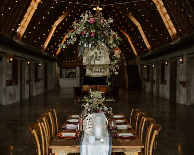 A long dining table is arranged under a decorated ceiling with string lights and floral hanging elements in a rustic venue.