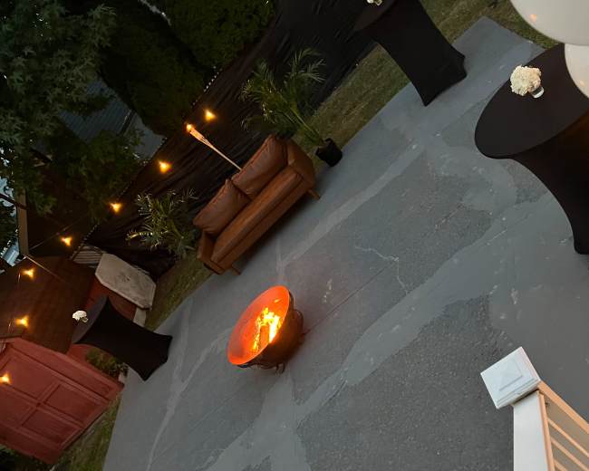 A patio area with a central fire pit surrounded by tall black tables and a brown couch, illuminated by string lights.