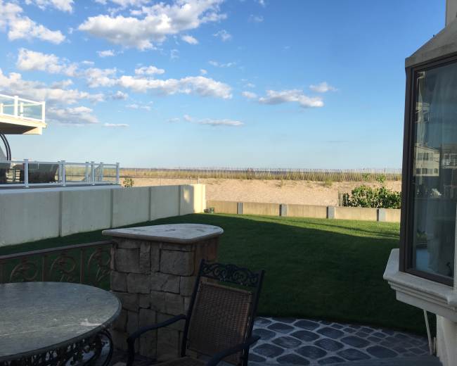 The image shows a backyard with a stone table and chair overlooking a sandy beach and grassy area under a partly cloudy sky.