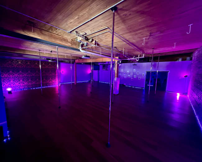 The image displays a spacious dance studio with polished wooden floors and multiple vertical poles, illuminated by purple and blue lighting.
