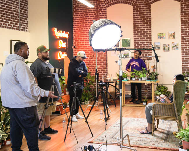 A group of four individuals sets up video equipment in a room filled with plants and brick walls.
