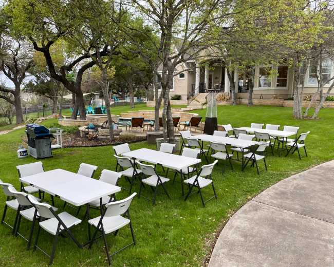 A set of white folding tables and chairs arranged on a grassy area near a house, with trees and a fire pit visible in the background.