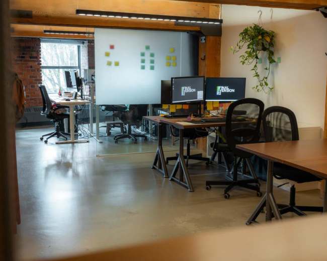 The image shows an office space with wooden beams, multiple desks, and computer monitors in a well-lit environment.