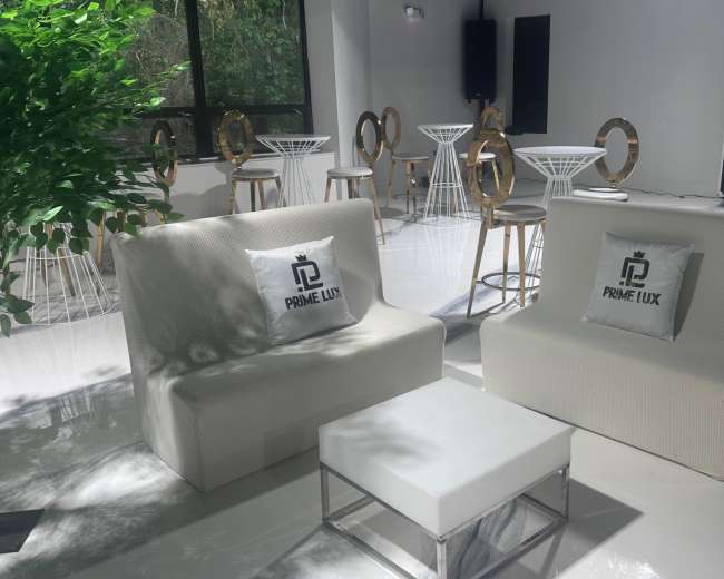 The image shows a modern, minimalist lounge area with white sofas and a coffee table, surrounded by high-top tables in a well-lit space.