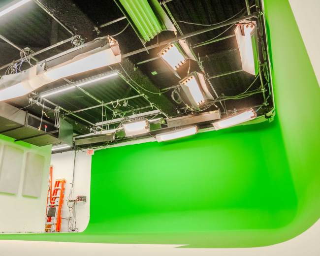 The image shows a studio with a green screen backdrop and overhead lighting fixtures.