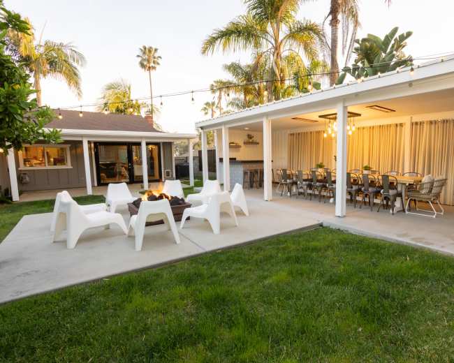 The image shows a backyard with a fire pit surrounded by white chairs, a covered patio with string lights, and a dining area with a long table.