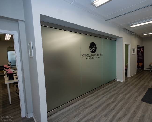The image shows a modern clinic interior with a frosted glass wall featuring the logo of "Advanced Aesthetics Skin Center" and a clean, minimalist design.