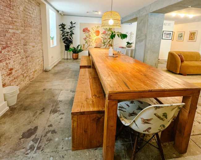 The image shows a spacious room with a large wooden dining table, surrounded by plants and featuring a decorative wall art.