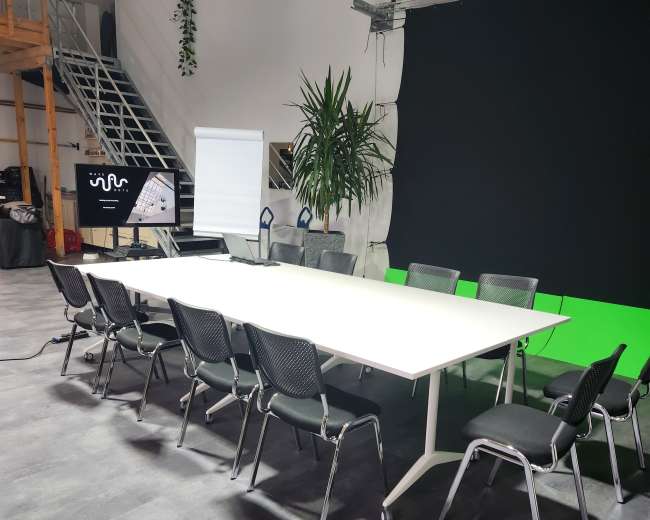 A large, white conference table surrounded by black mesh chairs is set up in a modern meeting room with a green screen backdrop.