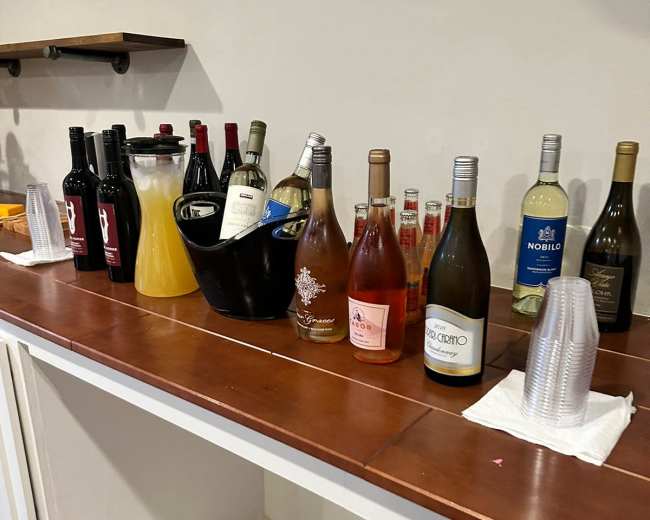 A variety of bottles, including wine and champagne, are arranged on a bar counter alongside plastic cups and a pitcher of juice.