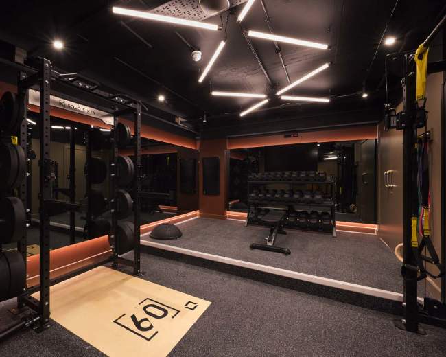 The image shows a modern gym interior equipped with weightlifting equipment, including a squat rack and dumbbell racks, along with a functional training area featuring a rubber floor and ceiling lights.