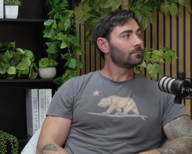 A man with a beard wearing a gray T-shirt sits next to a microphone against a backdrop of plants and shelves.