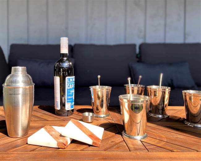 A metal cocktail shaker, several metallic cups, and two wooden coasters are arranged on a wooden table in front of a bottle of liquor and a dark sofa.