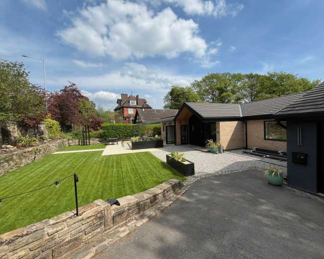 The image shows a modern, single-story house with a landscaped lawn, paved walkways, and a combination of stone and wood exterior finishes under a blue sky with some clouds.
