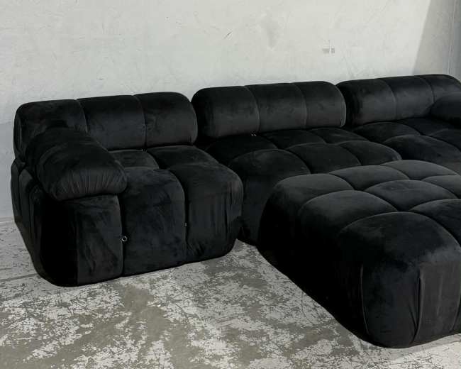 The image shows a modern black velvet sectional sofa with a plush, tufted design placed against a light-colored wall and textured floor.