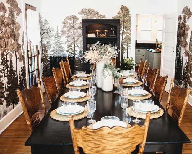 A large dining table is set for a meal, surrounded by wooden chairs and featuring wallpaper with a nature-inspired landscape.