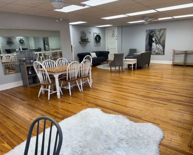 The image shows a spacious, well-lit room with wooden floors, featuring a dining table with white chairs, a grey sofa, various seating arrangements, and a decorative wall painting.