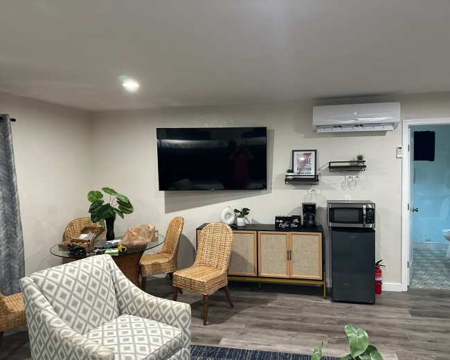 The living area features a round glass dining table, two wicker chairs, a sofa, a wall-mounted TV, and a kitchenette with a microwave and air conditioning unit.