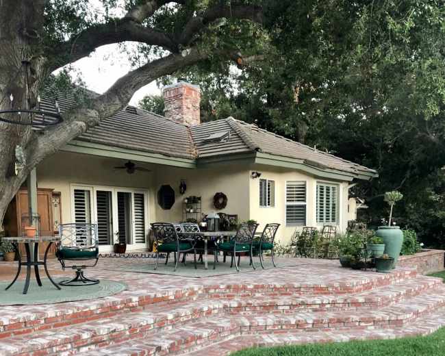 The image shows a patio area with brick steps leading up to a house surrounded by greenery and outdoor furniture.