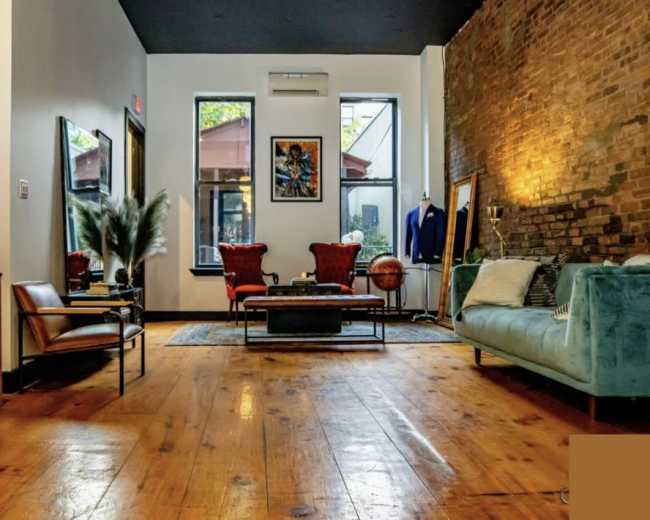 The image shows a well-furnished room with exposed brick walls, wooden flooring, and a mixture of seating options, including a teal couch and red armchairs.