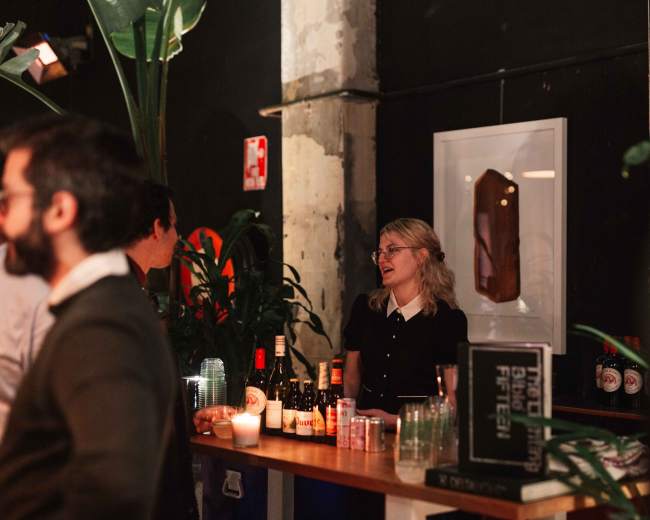 A bartender stands behind a wooden bar, surrounded by plants and bottles, engaging with customers in a dimly lit venue.