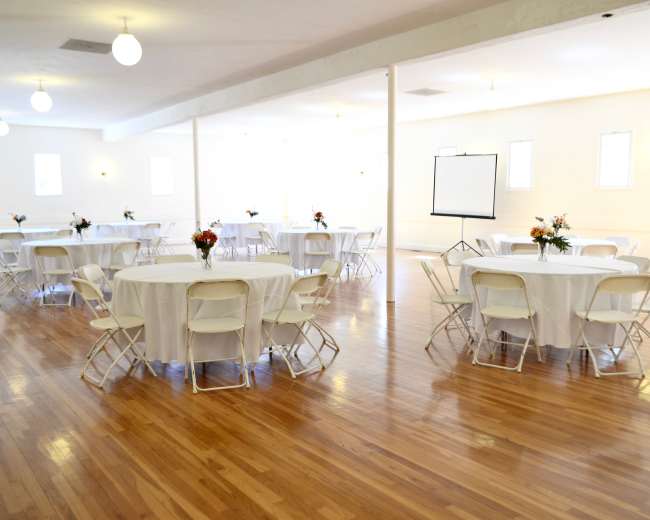The image shows a spacious reception hall with several round tables covered in white linens, surrounded by white folding chairs, and featuring a blank easel on one side.