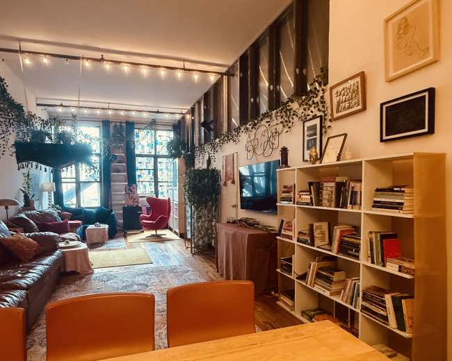 The image shows a cozy living space with a mix of seating options, shelves filled with books, and greenery adorned with string lights.