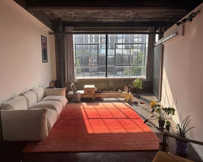 A spacious room with a large window, wooden beams, a white couch, orange rug, and plants near the sunlight.