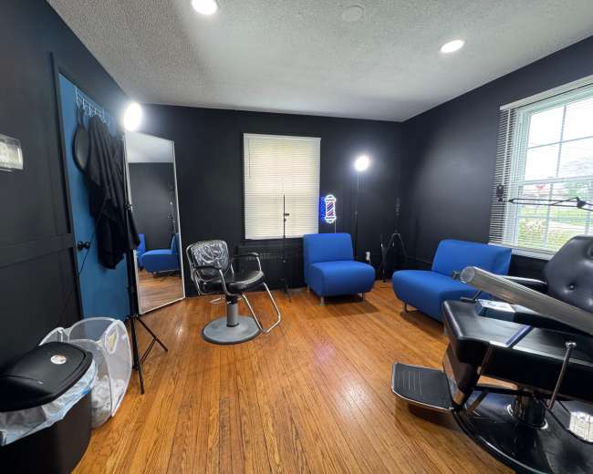 The image shows a barbershop interior with black walls, a chair for haircuts, a mirror, and two blue chairs.