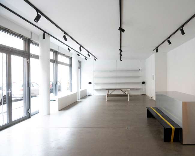The image shows a minimalist interior space with white walls, a long wooden table, a few shelves, and seating that features a yellow stripe.