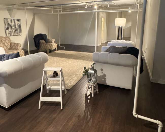The image shows a spacious basement living area with two sofas facing a central rug, along with a ladder and a light fixture illuminating the space.