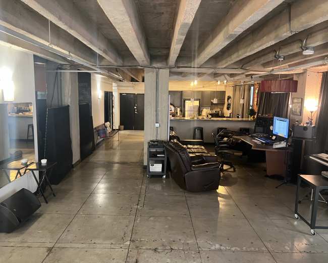 The image shows an open, spacious room with concrete floors, visible supports overhead, and areas designated for both living and working, including a kitchen and seating arrangement.