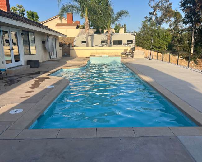 A rectangular swimming pool with clear blue water is surrounded by a stone deck and palm trees, with an RV parked nearby.