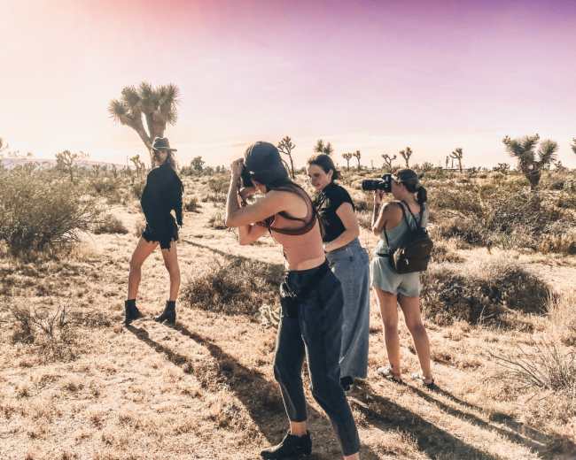 Four women are outdoors in a desert landscape, with Joshua trees in the background, as one models while the others take photographs.