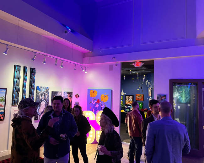 People socialize during an art exhibition featuring colorful artwork and vibrant lighting in a gallery space.