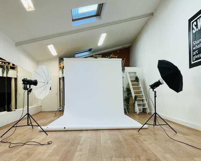 A photography studio features a white backdrop with lighting equipment set up on either side, along with a staircase and mirrors in the background.