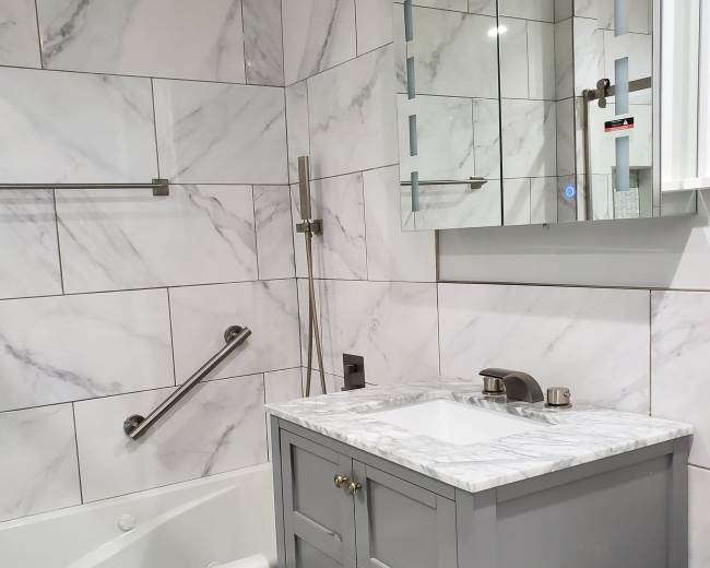 The image shows a modern bathroom featuring a bathtub, a marble vanity with two drawers, and a wall-mounted mirror above it, all surrounded by marble-tiled walls.