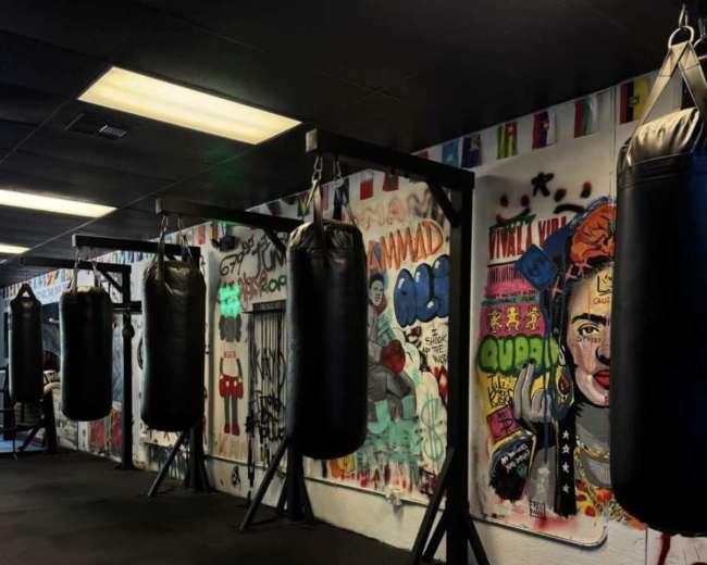 The image shows a gym interior featuring several black punching bags suspended from the ceiling, with colorful graffiti art covering the walls.