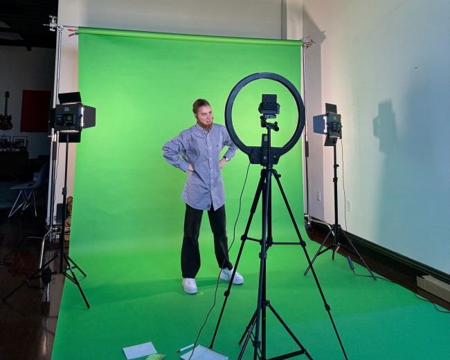 A person stands in front of a green screen setup, flanked by lights and a ring light.