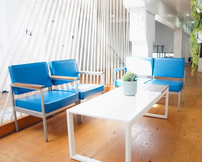 A bright, modern lounge area features blue chairs and a white table set on a wooden floor, illuminated by natural light.