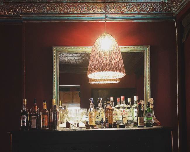 A well-lit bar area features a variety of liquor bottles arranged neatly on a dark wooden surface beneath a decorative mirror.