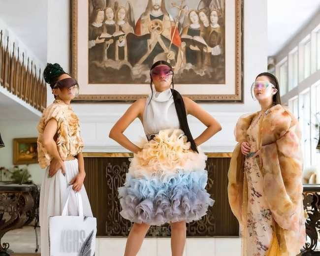 Three models pose in a spacious room with a large painting on the wall, each wearing unique fashion outfits and visors.