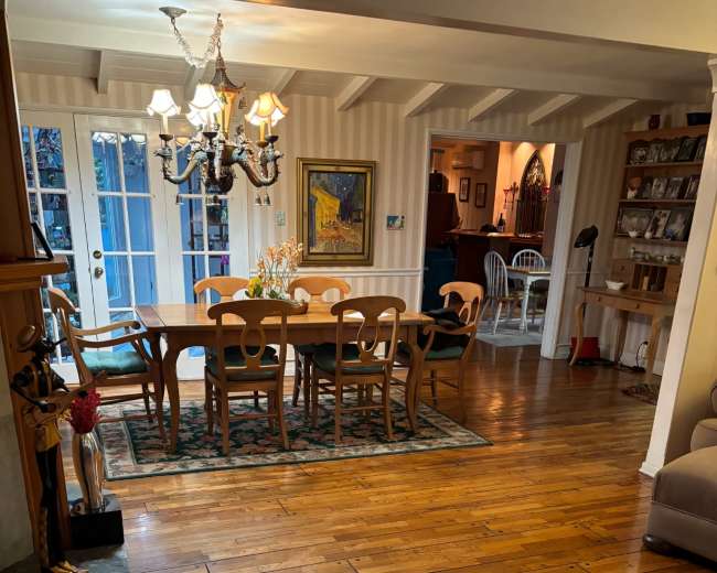 The image shows a dining area with a large wooden table surrounded by chairs, a chandelier overhead, and a view into a living space with warm wood flooring.
