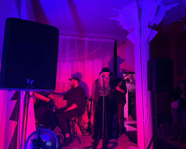 A singer stands on stage with a microphone while a musician plays a keyboard in a dimly lit venue illuminated by purple and pink lights.