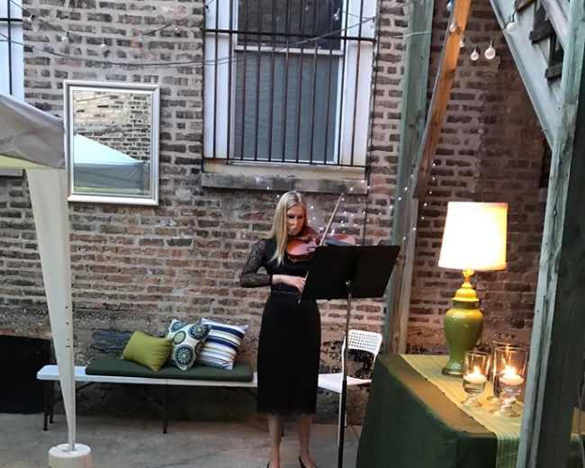A woman plays the violin in a courtyard setting, with a music stand and a table adorned with a lamp and decorative items nearby.