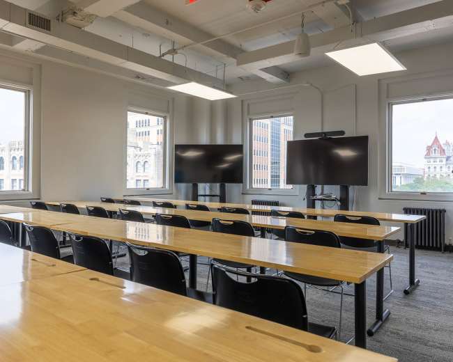 The image shows a modern classroom with rows of wooden tables and black chairs, featuring two large screens at the front and windows providing natural light.