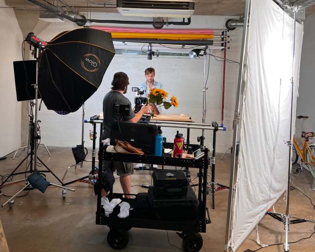 A photographer is setting up equipment and arranging flowers on a table in a studio with various lighting and a backdrop.