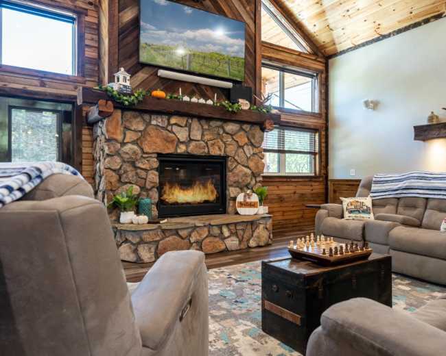 The image depicts a cozy living room featuring a stone fireplace, a mounted deer head, and a chess set on a small table, with wooden walls and a television mounted above the fireplace.