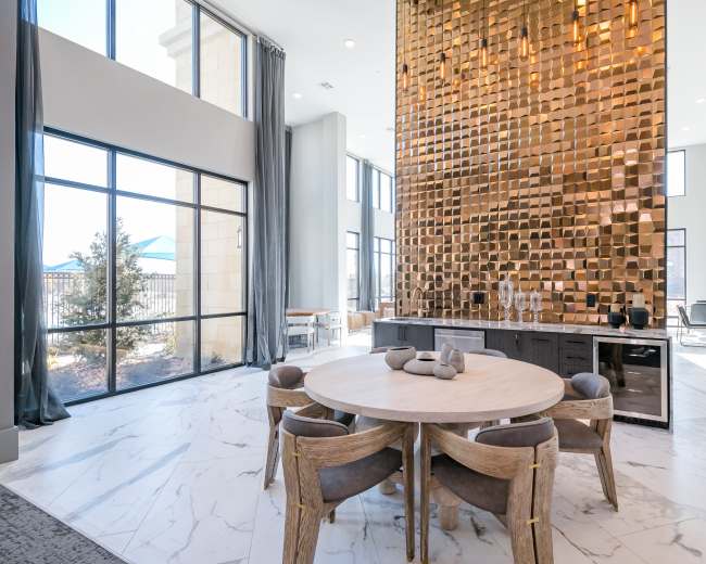 A spacious modern room features a large round wooden table surrounded by upholstered chairs, with a striking gold-accented wall and large windows letting in natural light.